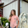 Woman in pink shirt stands outdoors by rustic building.